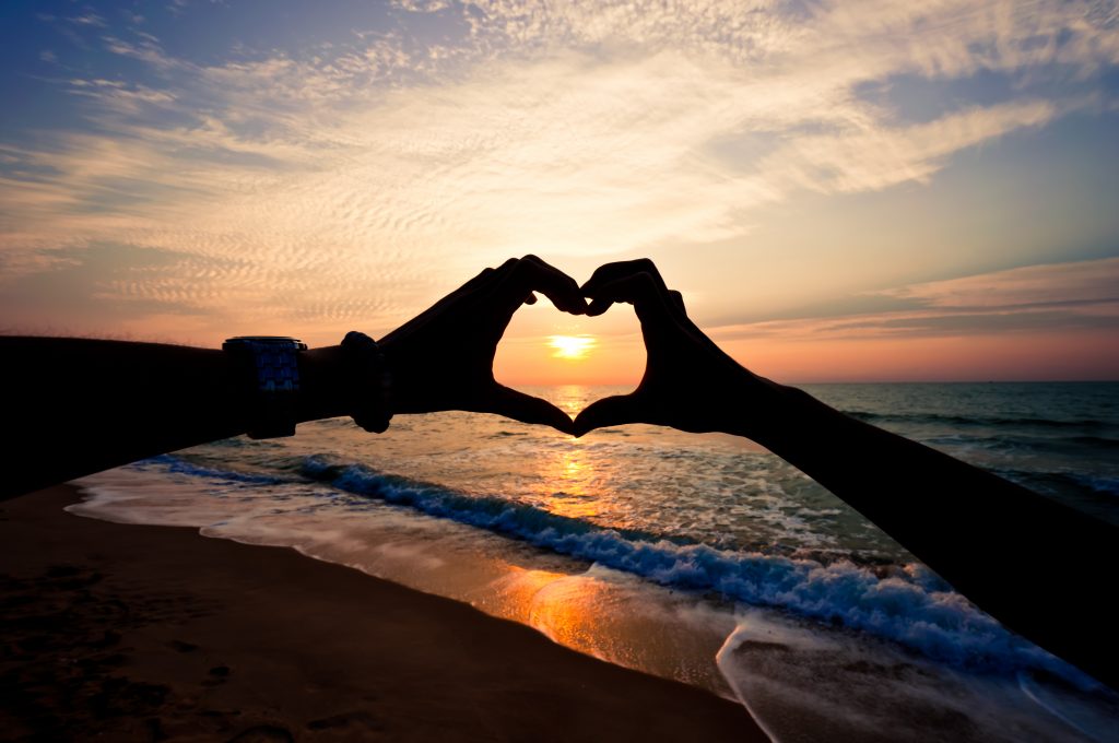 Silhouette hand in heart shape with sunrise in the middle and beach background
